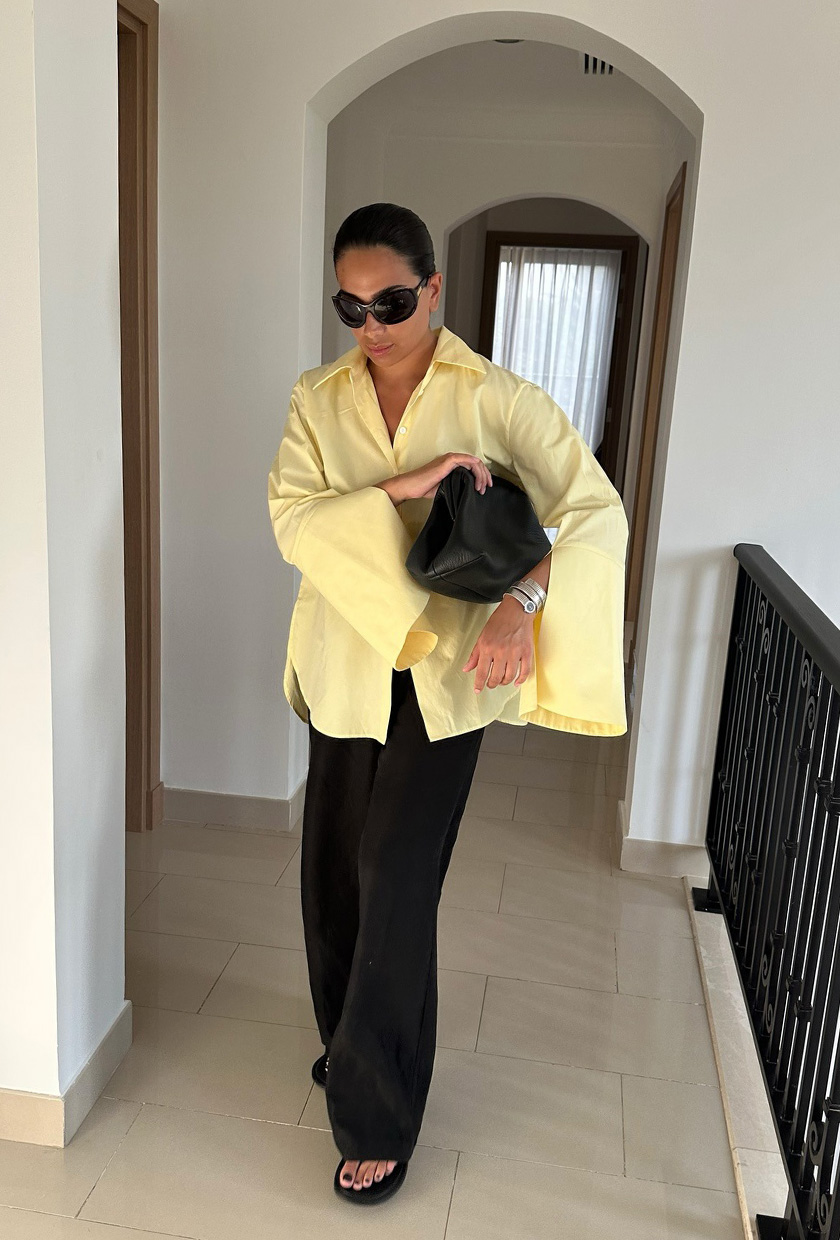 How to pack for Italy is shown in a picture of a woman wearing black sunglasses, an oversized yellow button down shirt, a black clutch bag, black trousers, and black heeled thong sandals