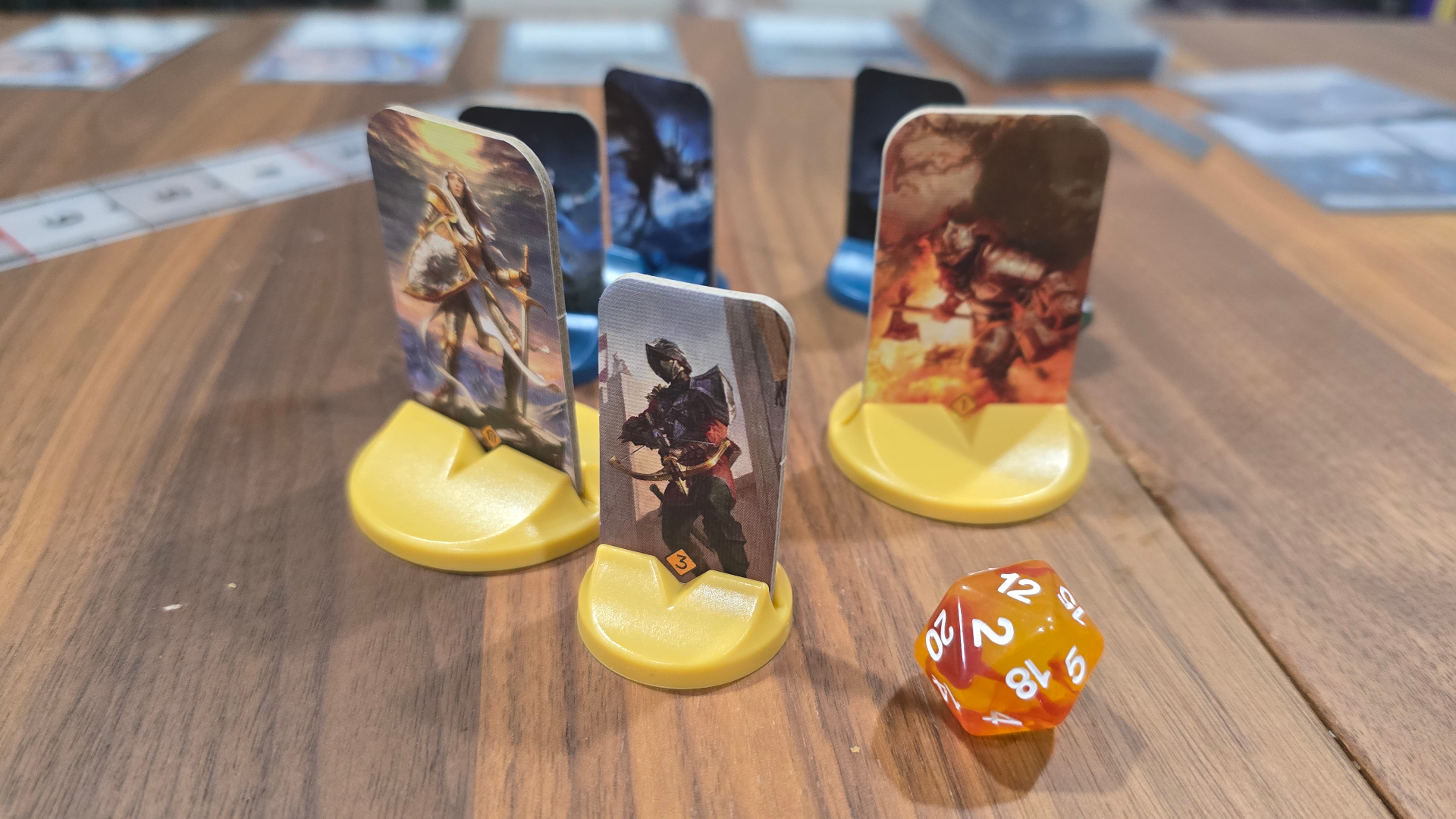 Malediction cardboard standees on a wooden table beside a 20-sided die