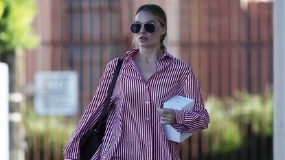 Margot Robbie wearing an oversized red and white button down with baggy blue jeans, sunglasses, and a black shoulder bag