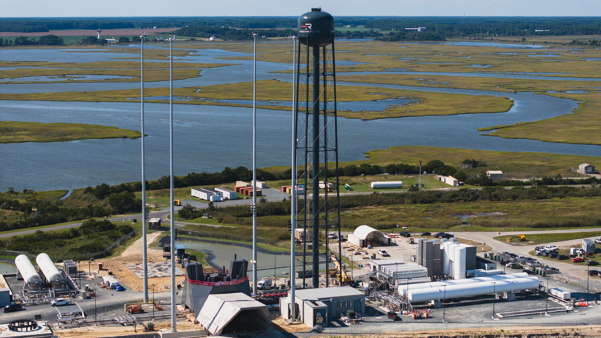 A zoomed out image of a the Rocket Lab launch complex, with a tall black water tower overlooking several buildings next to a wetland.