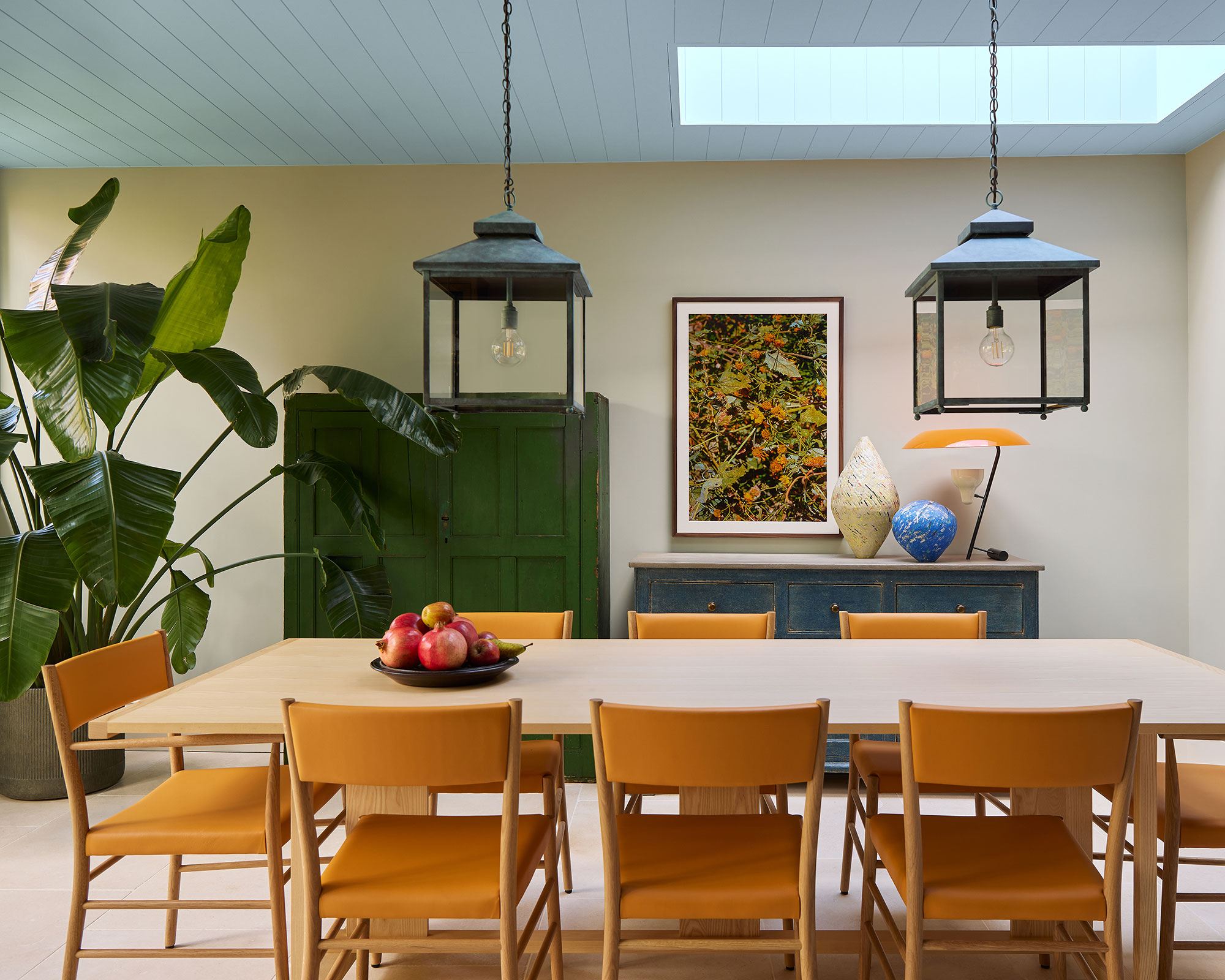An elegant dining area designed by Tom Morris at Morris Studio, featuring a light wood table surrounded by tan leather chairs. The space is characterized by a soft sky-blue paneled ceiling with a large skylight and a statement green armoire positioned next to a tall Bird of Paradise plant. Modern lantern pendants hang above the table, while a blue sideboard and organic art pieces add layers of sophisticated color to the neutral walls.