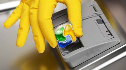 Close-up of hand in yellow rubber glove putting detergent tablet into dishwasher