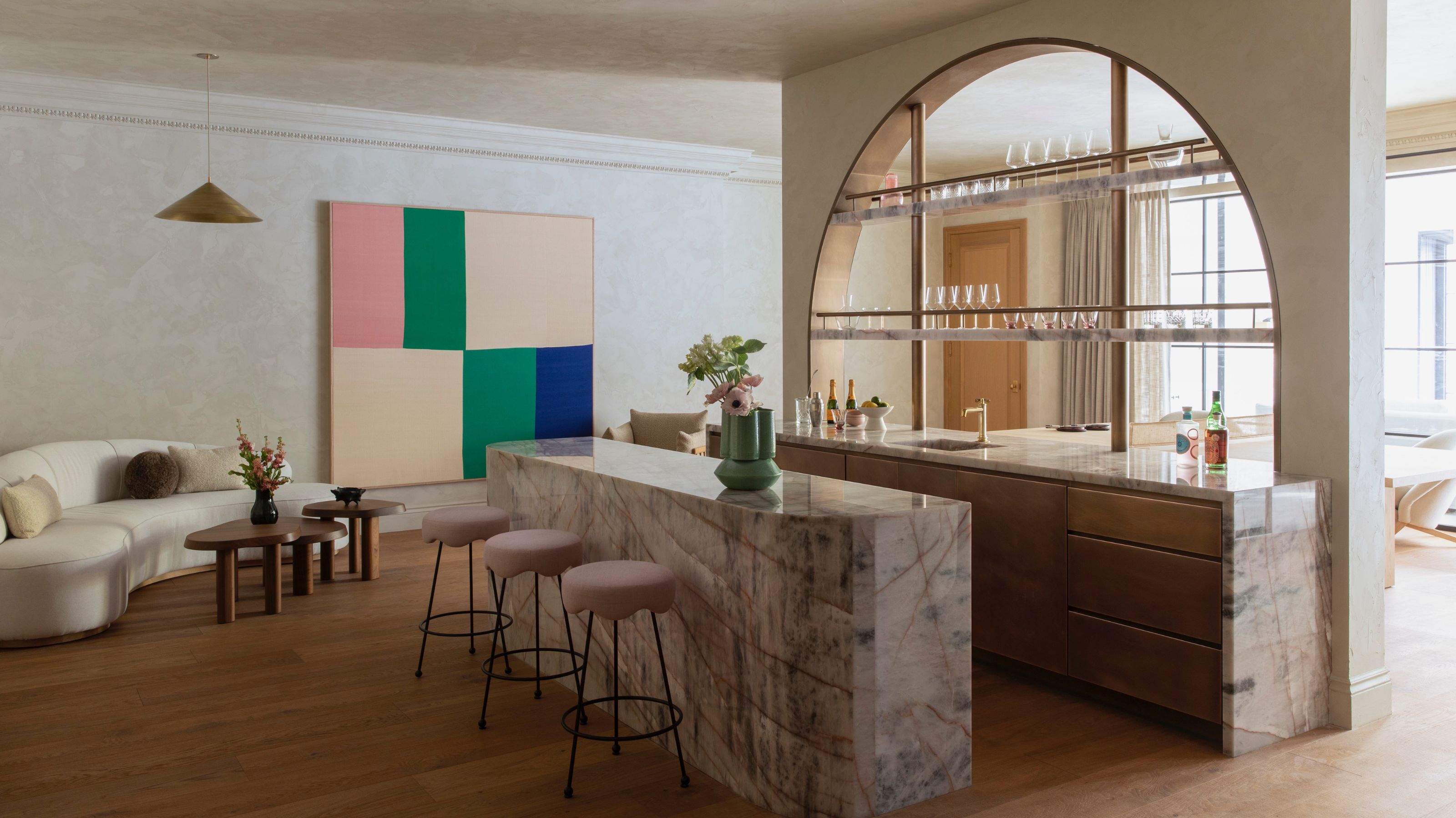 A luxurious open-concept lounge area featuring a dramatic, purple-veined marble waterfall island with pink scalloped barstools. The background includes a large arched architectural opening with glass shelves, a curved cream sofa, and vibrant abstract wall art