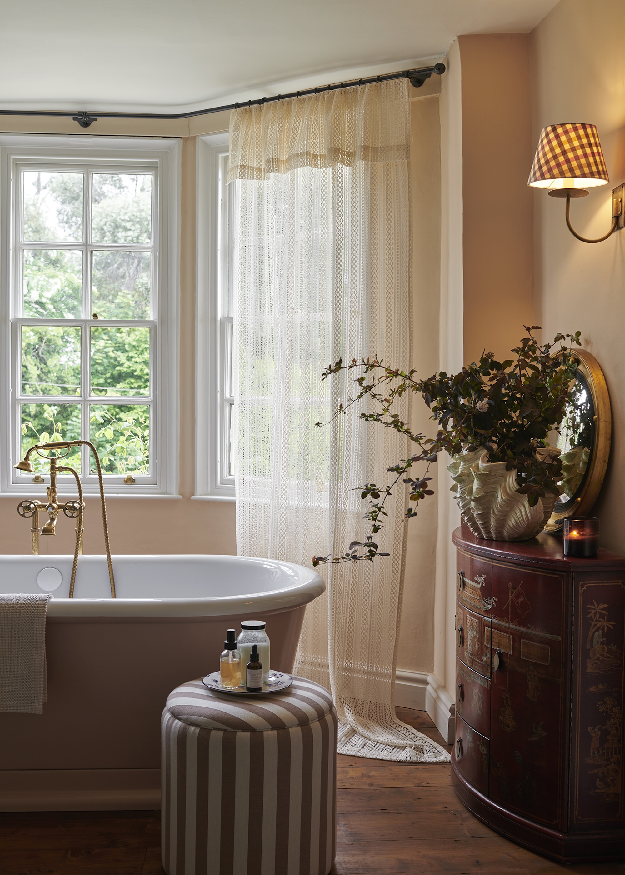 A whimsical style bathroom with full length lace curtains, a roll top bath, antique side board, a large vessel of foliage and striped pouffe