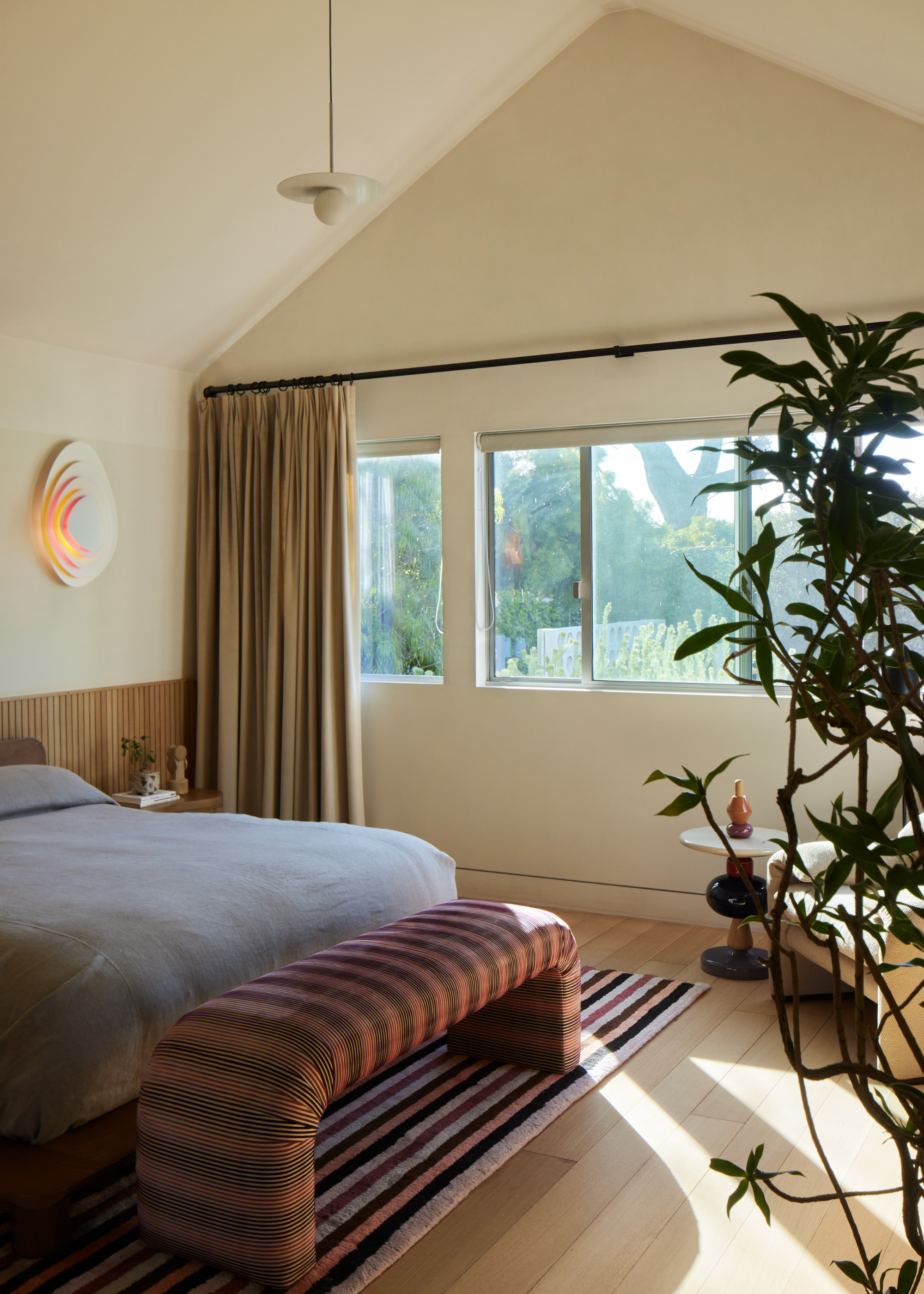 An off-white bedroom with a vaulted ceiling, large sunny windows, a houseplant, a sofa seat in the corner with a side table, a striped rug with a striped stool on top and a gray bed with a wooden headboard and a layered wall sconce