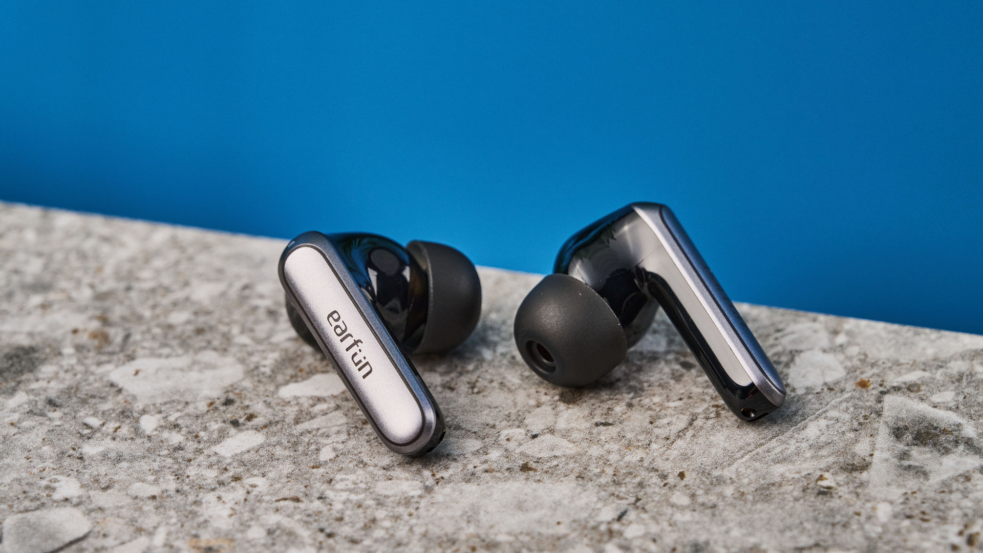 a pair of black and silver earbuds with silicone tips and a black charging case are photographed against a blue background