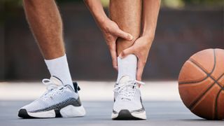 Bottom half of a basketball players legs are shown. He is wearing white socks and white trainers and is holding on to his left ankle as if in pain. There is an orange basketball next to his left foot.