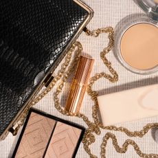 Bird's eye view of a black clutch bag, four make-up products and a brush and a bottle of perfume laid out in a flatlay against a cream background