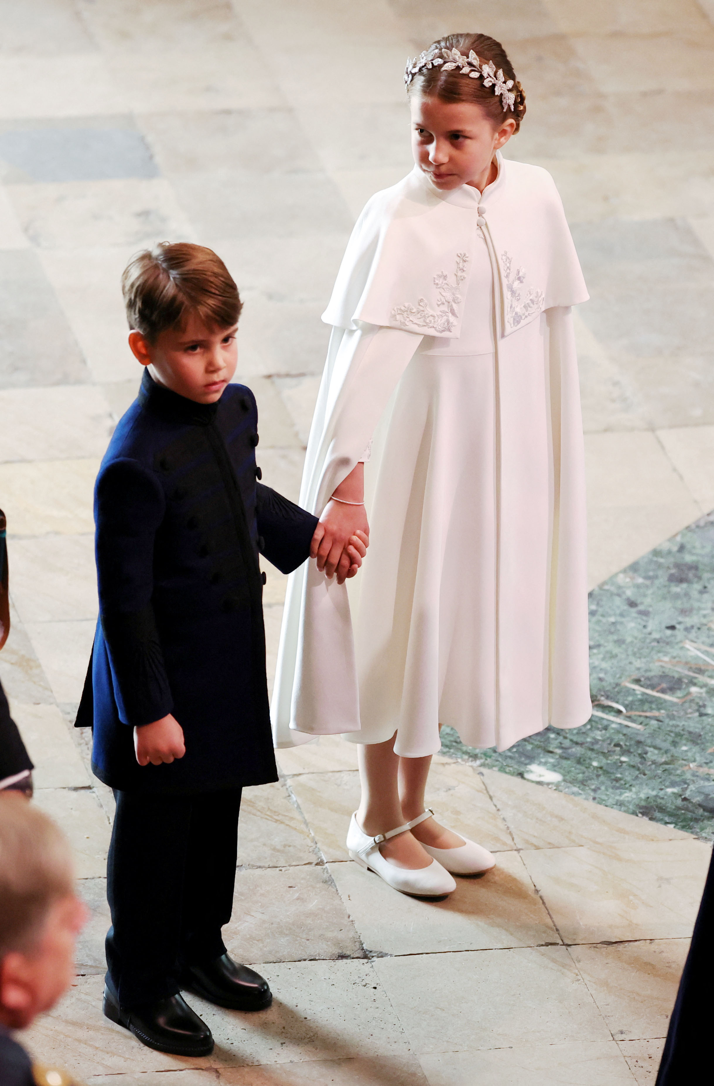 Princess Charlotte wearing a white cape holding hands with Prince Louis at King Charles's coronation