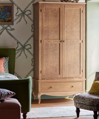 Freestanding pine two-door closet with brass hardware, green velvet bed, geometric rug, blush pink ottoman at the foot of the bed and colorful armchair and pillows. The wallpaper is cream with sage green large bows