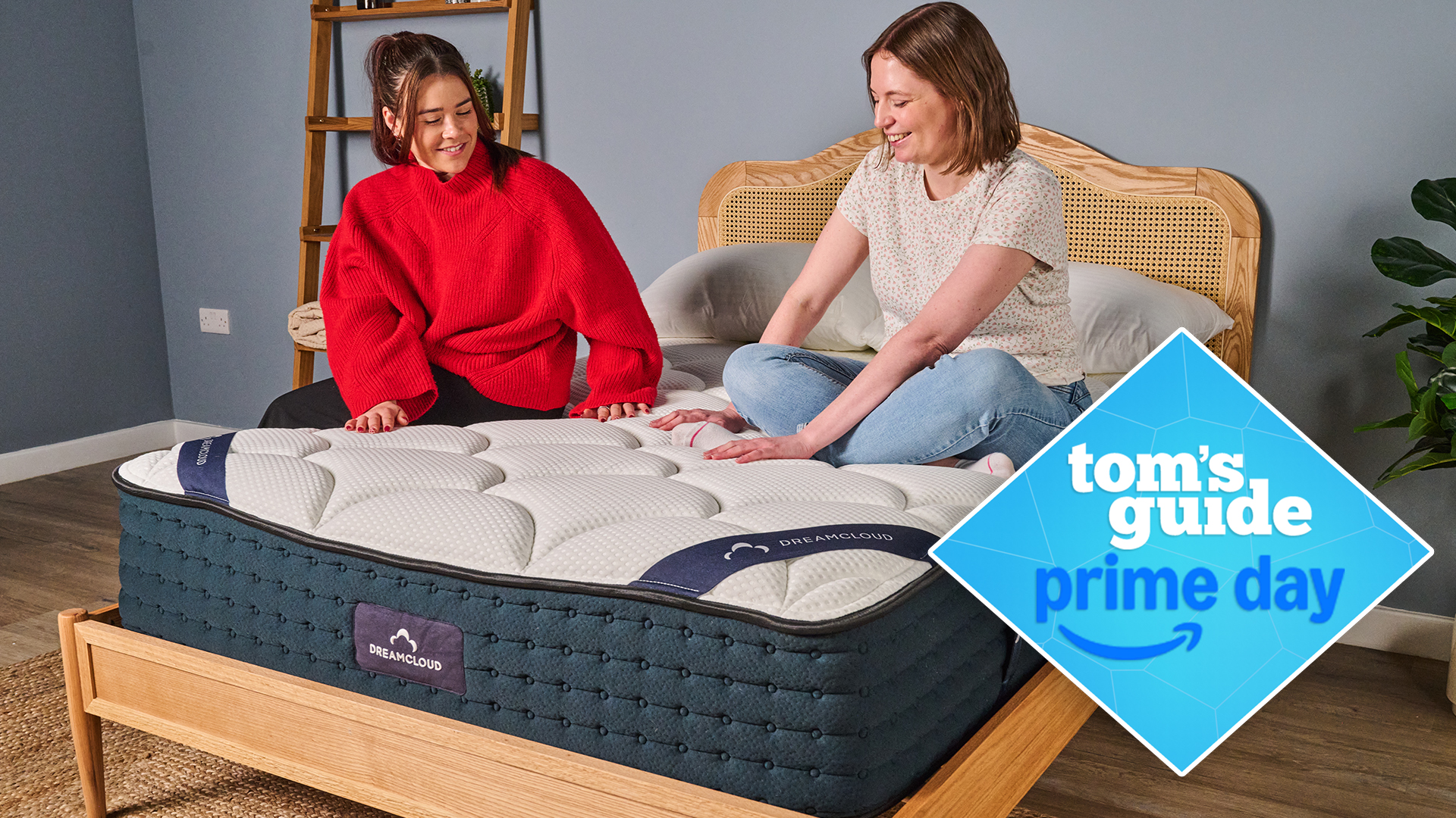 Two women sit on the DreamCloud Classic Hybrid mattress on a bed frame in a bedroom, a Tom&#039;s Guide Prime Day deals graphic in the bottom right corner