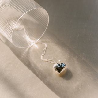 A silver heart locket on a long silver chain sits on a piece of white linen fabric