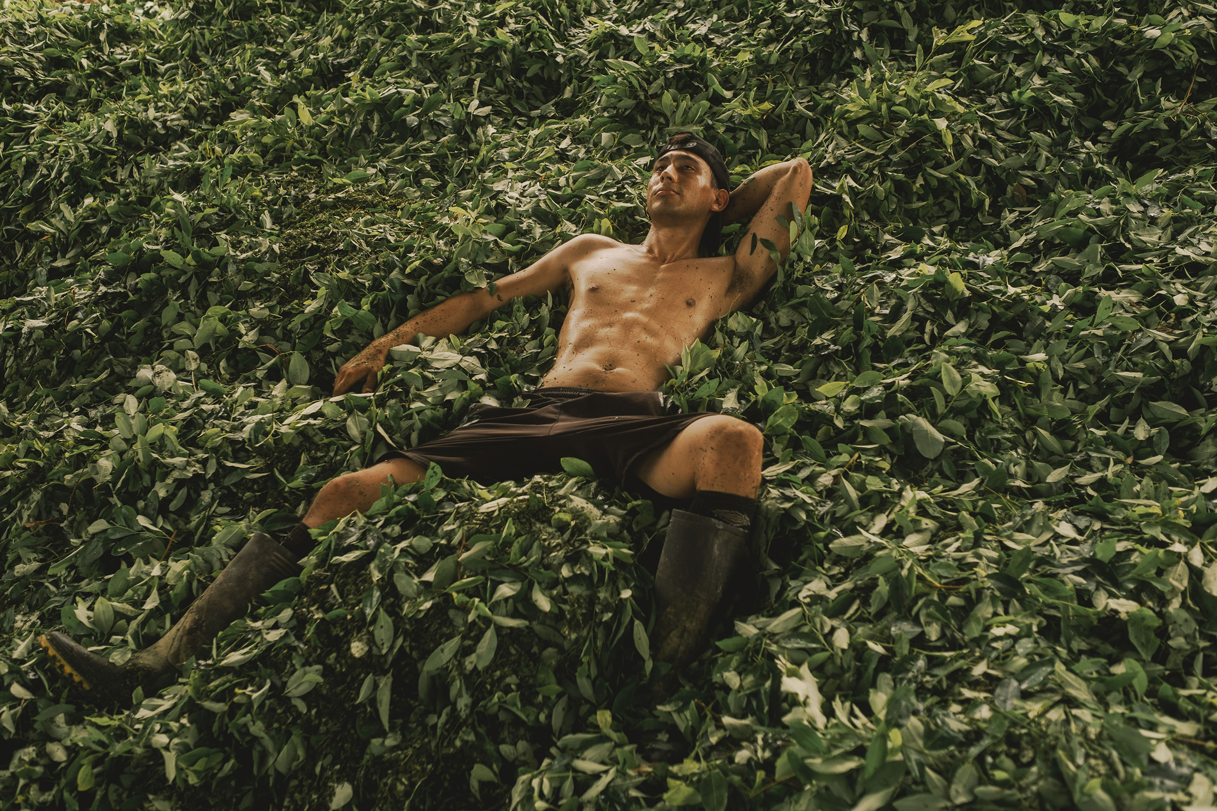A shirtless man in shorts and boots relaxes atop a large pile of green leaves, arms behind his head