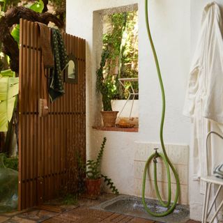 outdoor shower with Ikea screen, alcove in wall, watering can and hose shower, plants, towels, small mirror, tiled floor