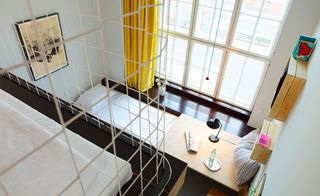 Hotel room, large white frame window, yellow curtains, white walls , grey floor, desk top with desk lamp, bottle of water and glass, striped pillow, on top of a single bed and mattress, wooden box shelves on wall, picture frame, metal cage with a mattress inside in the front left
