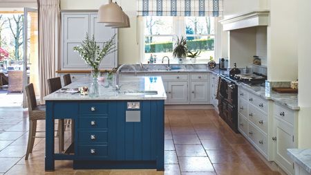 Bright kitchen with white cabinets, marble countertops, terracotta tiled flooring, and a blue kitchen island with large windows behind.
