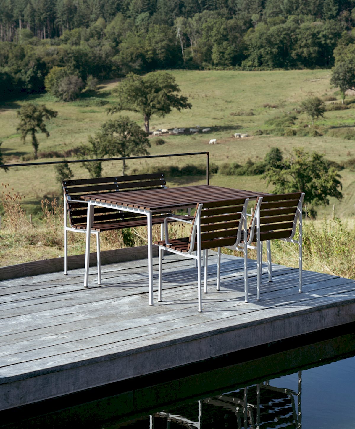 a wooden and aluminium table and chairs sits on a wooden deck by a pool overlooking green countryside