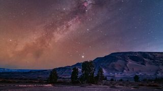 The glowing band of the Milky Way's galactic plane is pictured tumbling diagonally through a starry night sky above a mountainous region.