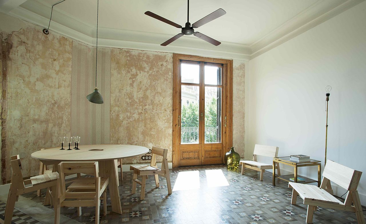 Interior view of a dining and seating area at Yök Casa + Cultura featuring a white ceiling with a fan and long pendant light and white and rustic walls. There is a round light wood dining table with glasses, light wood chairs, a side table with books and a floor lamp. The flooring is multicoloured and the tall glass doors have a wooden frame