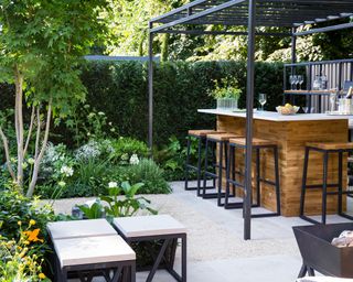 Sleek, modern garden bar ideas in a pale paved patio garden with square flower beds, under a square black metal pergola.