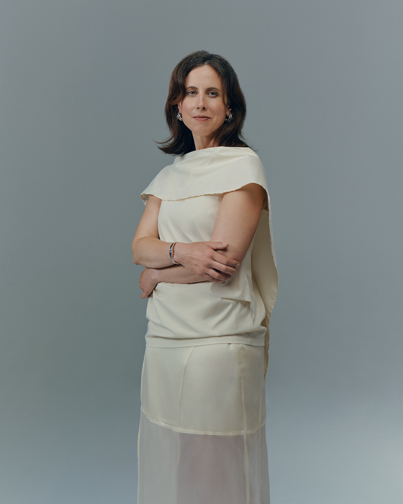 Portrait of a woman in an ivory outfit standing against a plain gray background with her arms crossed.