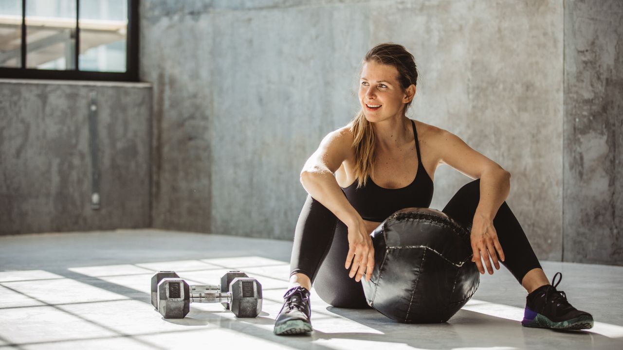 A woman sits in an empty room on the floor, with her legs bent and her arms resting on her knees. In front of her is a medicine ball and to her side is a set of dumbbells.