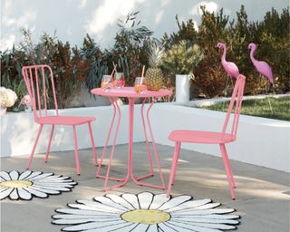 Heidi Outdoor Bistro Set in pink outside in yard with white walls and two daisy rugs