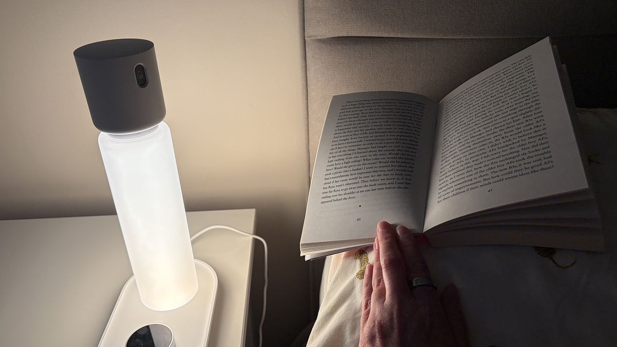 The Sleepal AI Lamp on a nightstand in our reviewer's bedroom. The lamp is lit up and is throwing light on a book, placed on the pillow next to the lamp. The book is being held open by the reviewer's hand