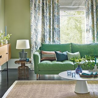 Green living room with sage walls and an emerald sofa in front of a large window lined with green and blue botanical print curtains