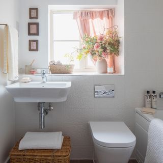 a compact bathroom with basi and loo, small window with a pink striped curtain and a large basket for storage