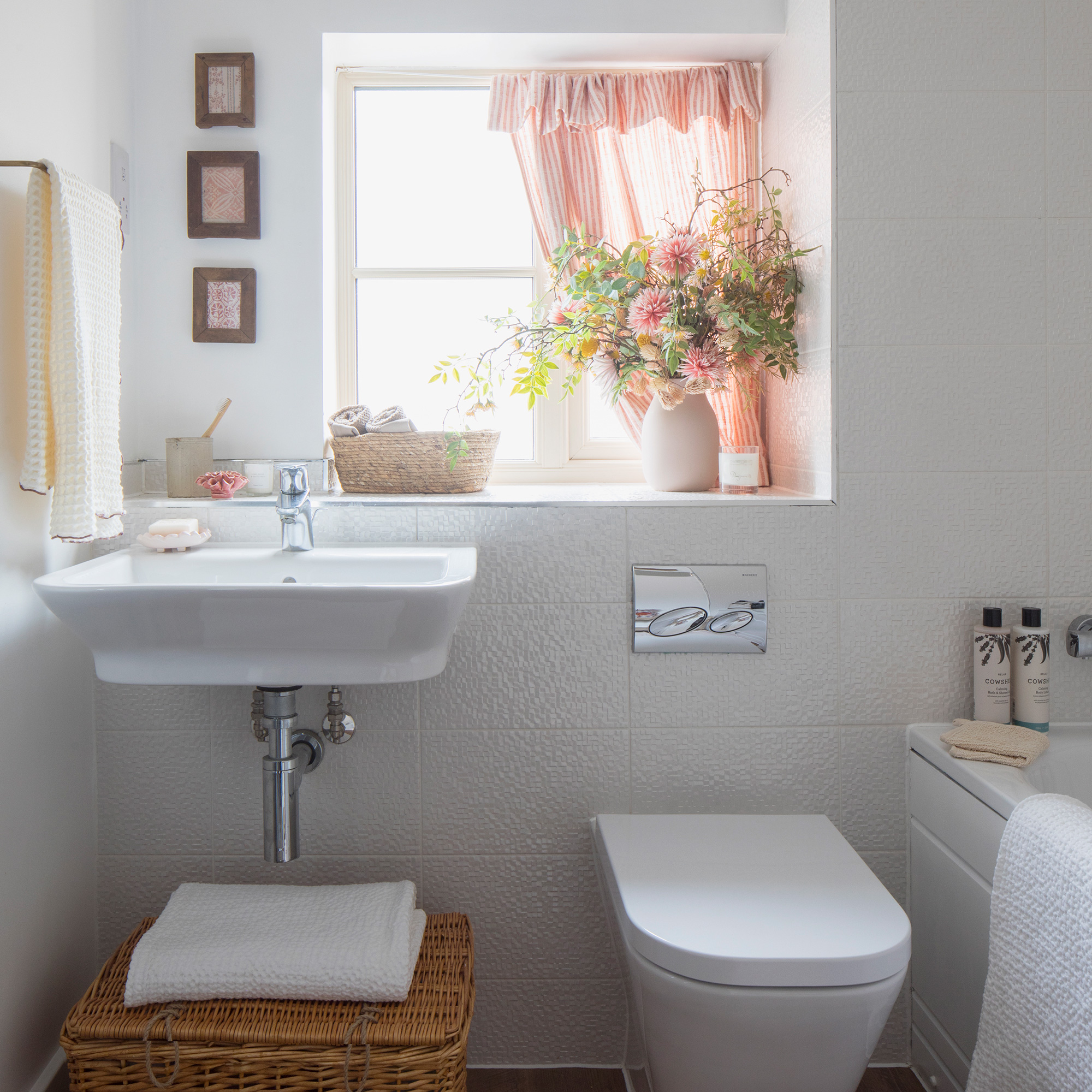 Un baño compacto con basi y loo, ventana pequeña con una cortina rosa a rayas y una canasta grande para el almacenamiento