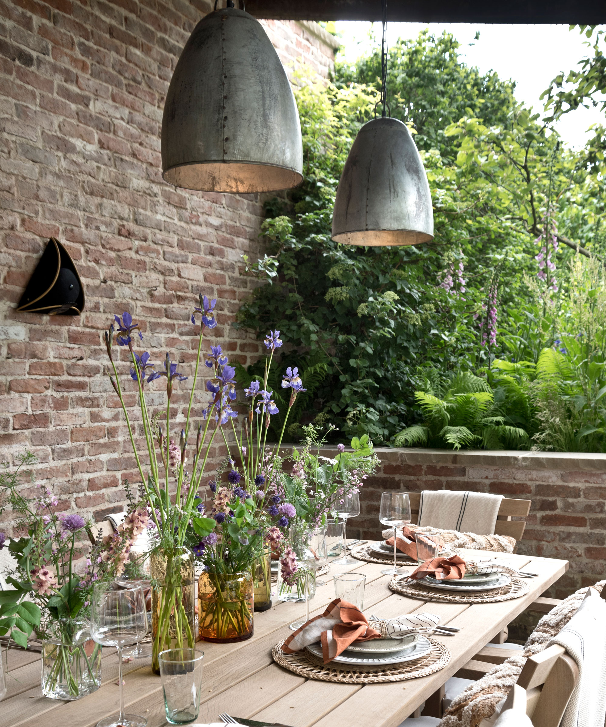 outdoor dining area laid with china, glassware and napkins, with overhead outdoor pendant lighting, enclosed by a solid brick wall and a low brick wall, with trees and shrubs beyond