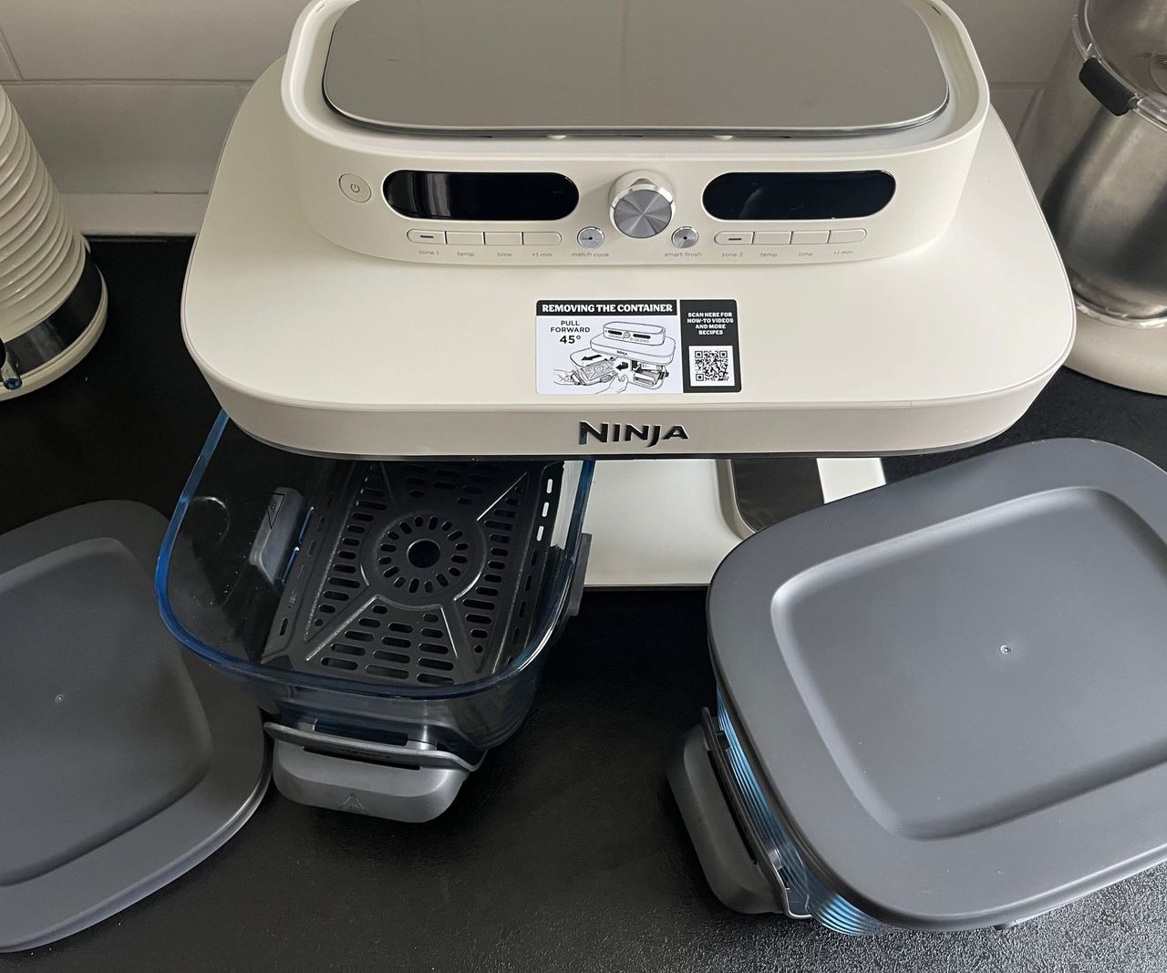 A ninja Crispi DualZone air fryer on a black kitchen counter with the instructions and two grey lids.