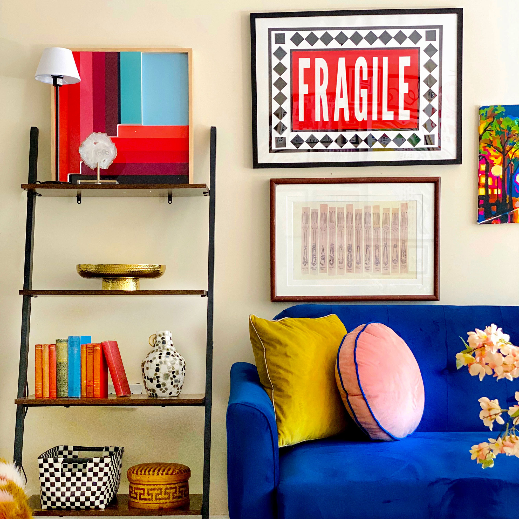 Blue sofa in living room with pink and yellow cushions below artwork on wall and beside open shelving unit holding books, vases and baskets
