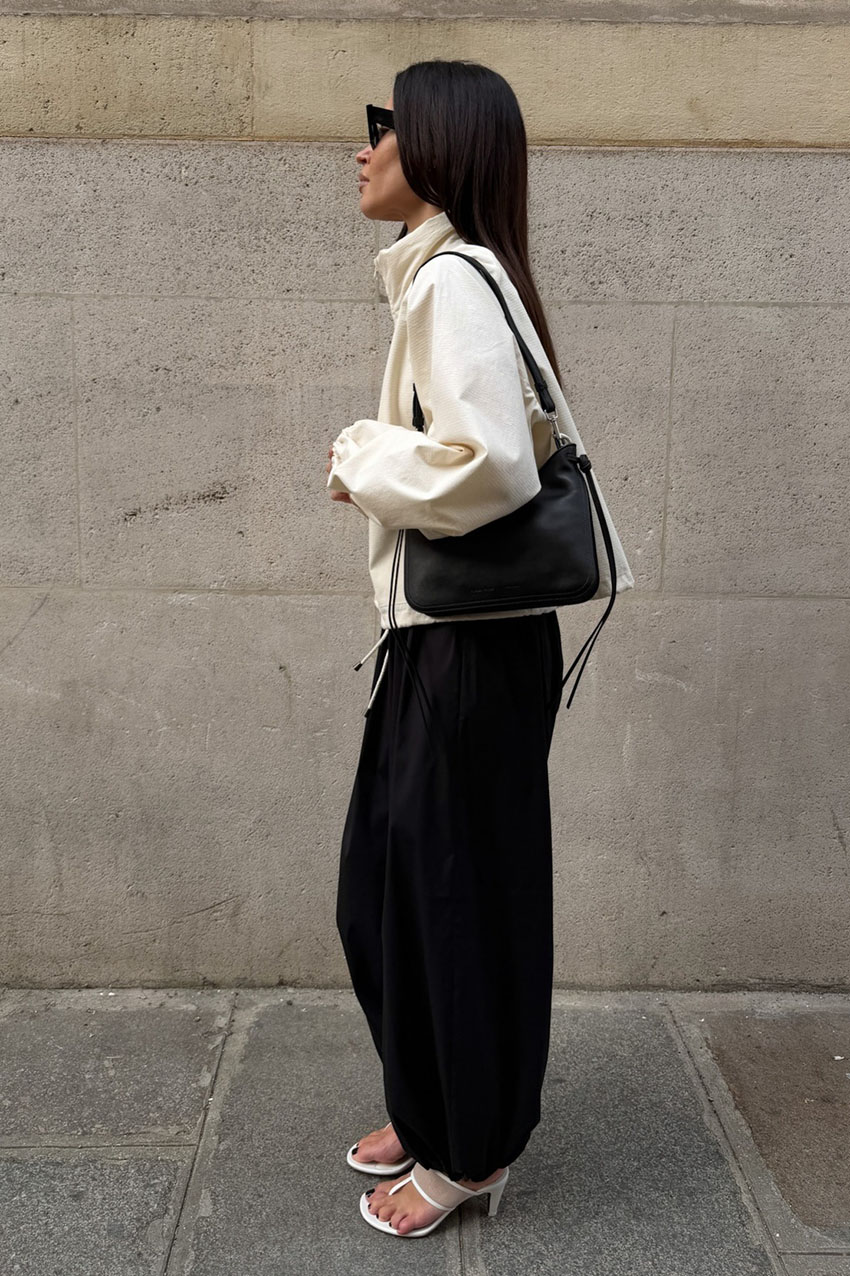 A woman wearing a white funnel-neck jacket, black balloon pants, white kitten heel thong sandals, and a black bag.
