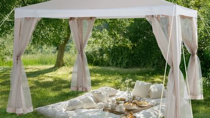 Beige soft-top gazebo with mesh standing over a charming picnic scene on a grassy lawn surrounded by trees