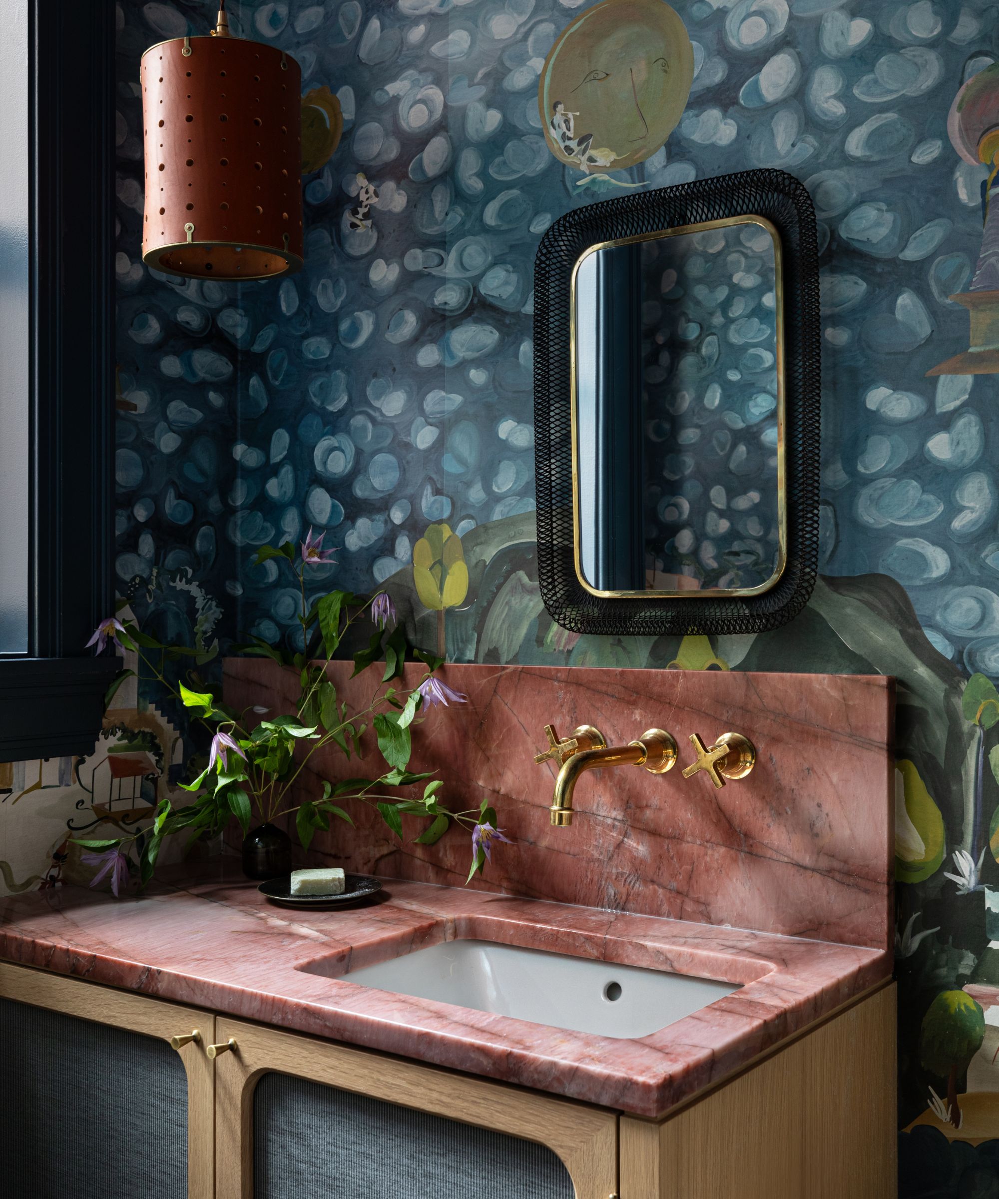 powder room with blue patterned wallpaper on the walls with a mirror and pink quartz countertop on wooden vanity and brass hardware