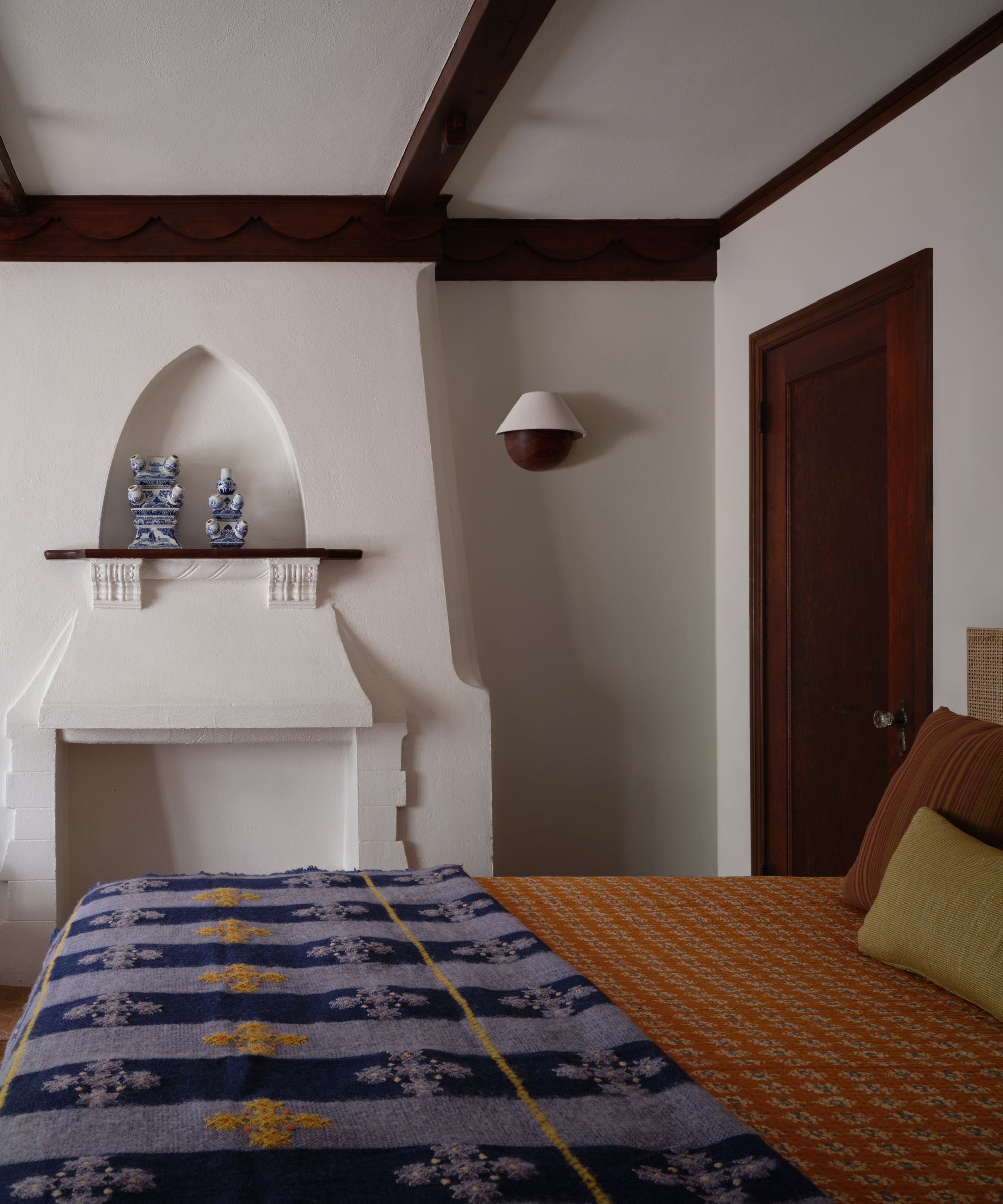 A bedroom view showing an architectural white fireplace with a pointed niche above, a blue patterned blanket on the bed, and dark wood ceiling trim