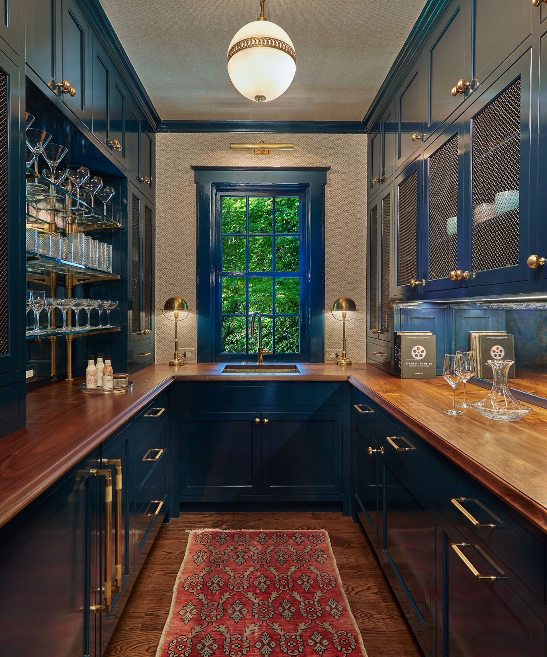 A kitchen with dark blue cabinets, wooden countertops, gold hardware, and a dark wood floor topped with a red Persian runner rug