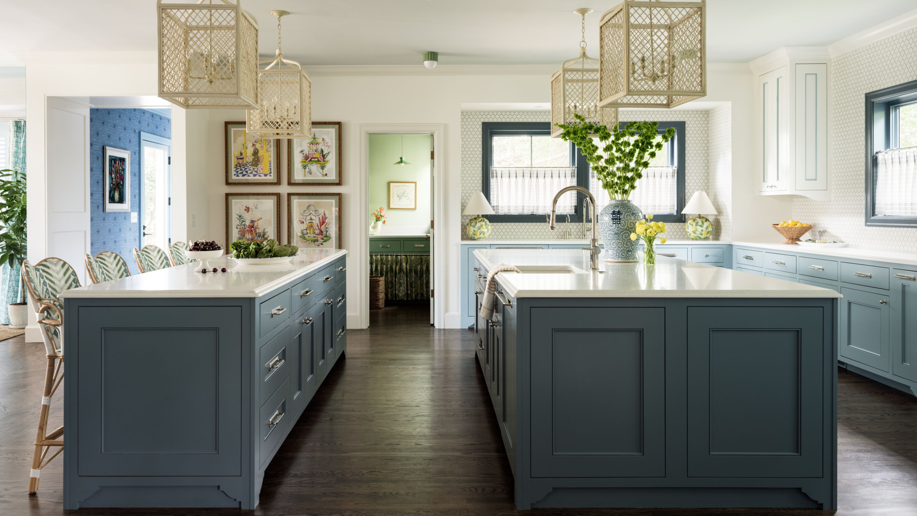 a light kitchen with subtle wallpaper details light blue cabinetry and double islands with cage lights and a view through to a green pantry