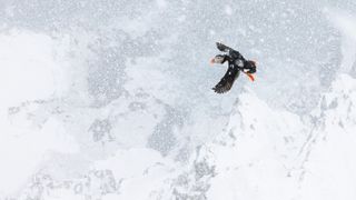 A puffin with a bright orange beak and feet soars through the snowy sky, wings extended wide. The wintery backdrop adds a serene and chilly atmosphere