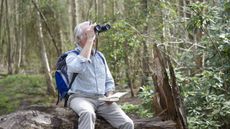 An older man on a hike uses binoculars while holding a guide book.