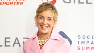 Portrait of Sharon Stone smiling, with a blonde pixie cut and wearing a pink trench coat and pearl necklace