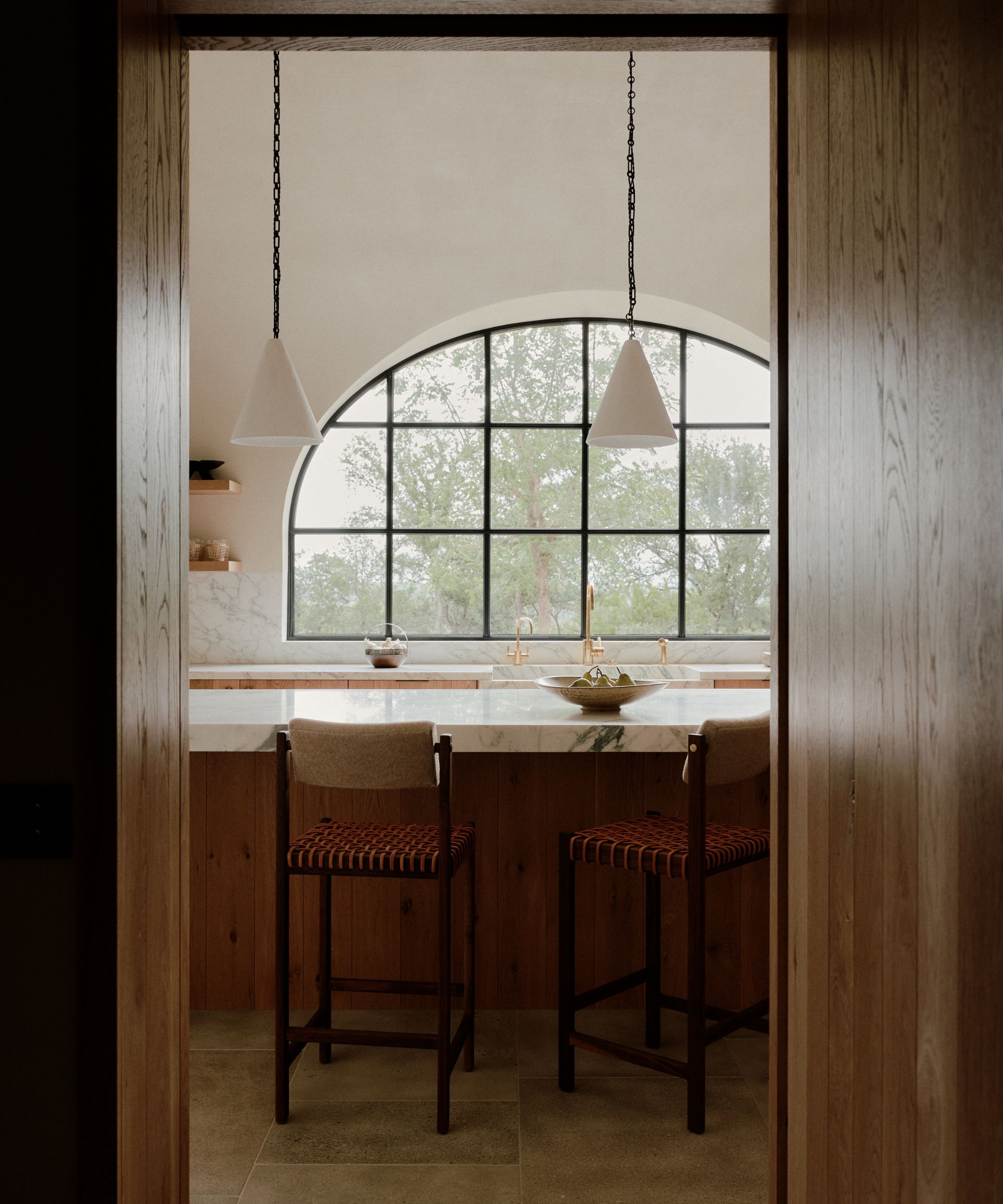 A view into the kitchen framed by wooden doors, centering on the marble island and a large arched window overlooking green trees. Two white pendant lights hang symmetrically over the island