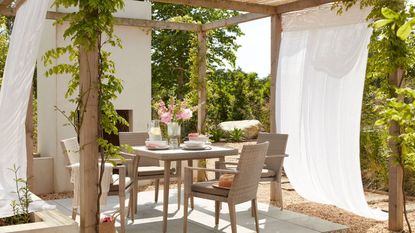 A pergola with outdoor curtains hanging from the rafters over an outdoor dining table