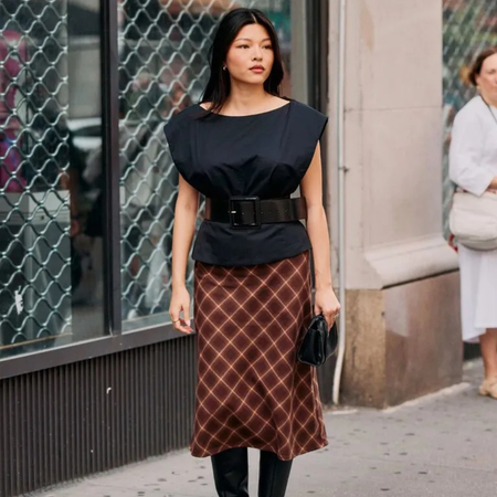 A NYFW S/S 26 attendee wears a black boat neck top with chunky waist belt, red check midi skirt, black leather boots and a leather clutch bag
