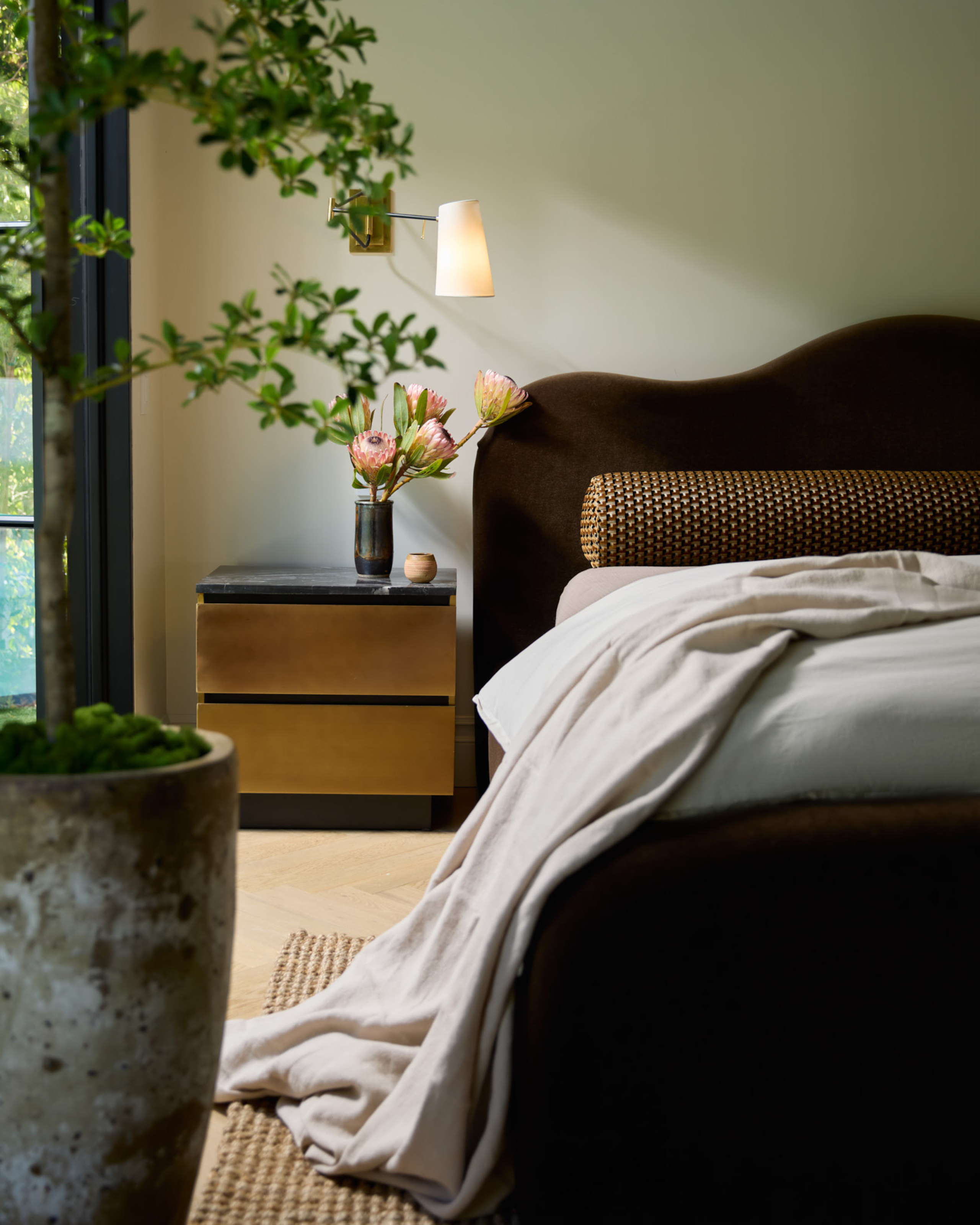 A bedroom with soft green walls, brown velvet wavy bed, brown patterned bolster cushion, brown and black side table and beige throw