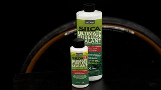 Two white bottles with green labesl stand in front of a tanwall gravel tyre against a black background