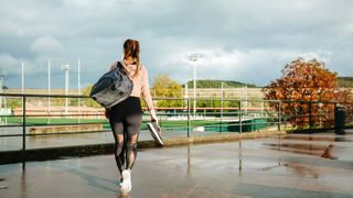 Woman walking towards sports fields carrying bag slung over shoulder and trainers