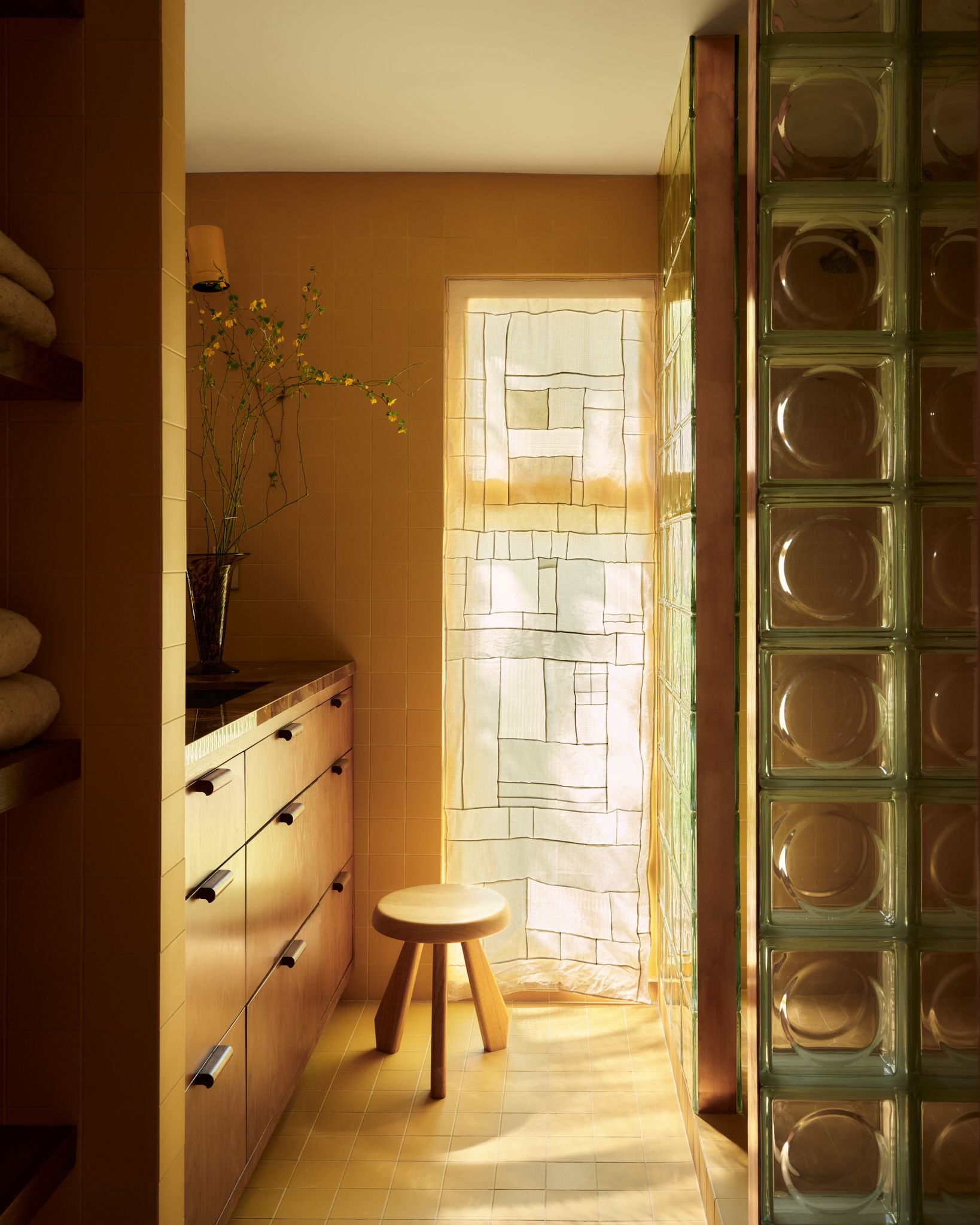 A modern bathroom with a patterned sheer curtain next to a stool and built-in vanity unit with green-framed glass blocks on the opposite side of the space